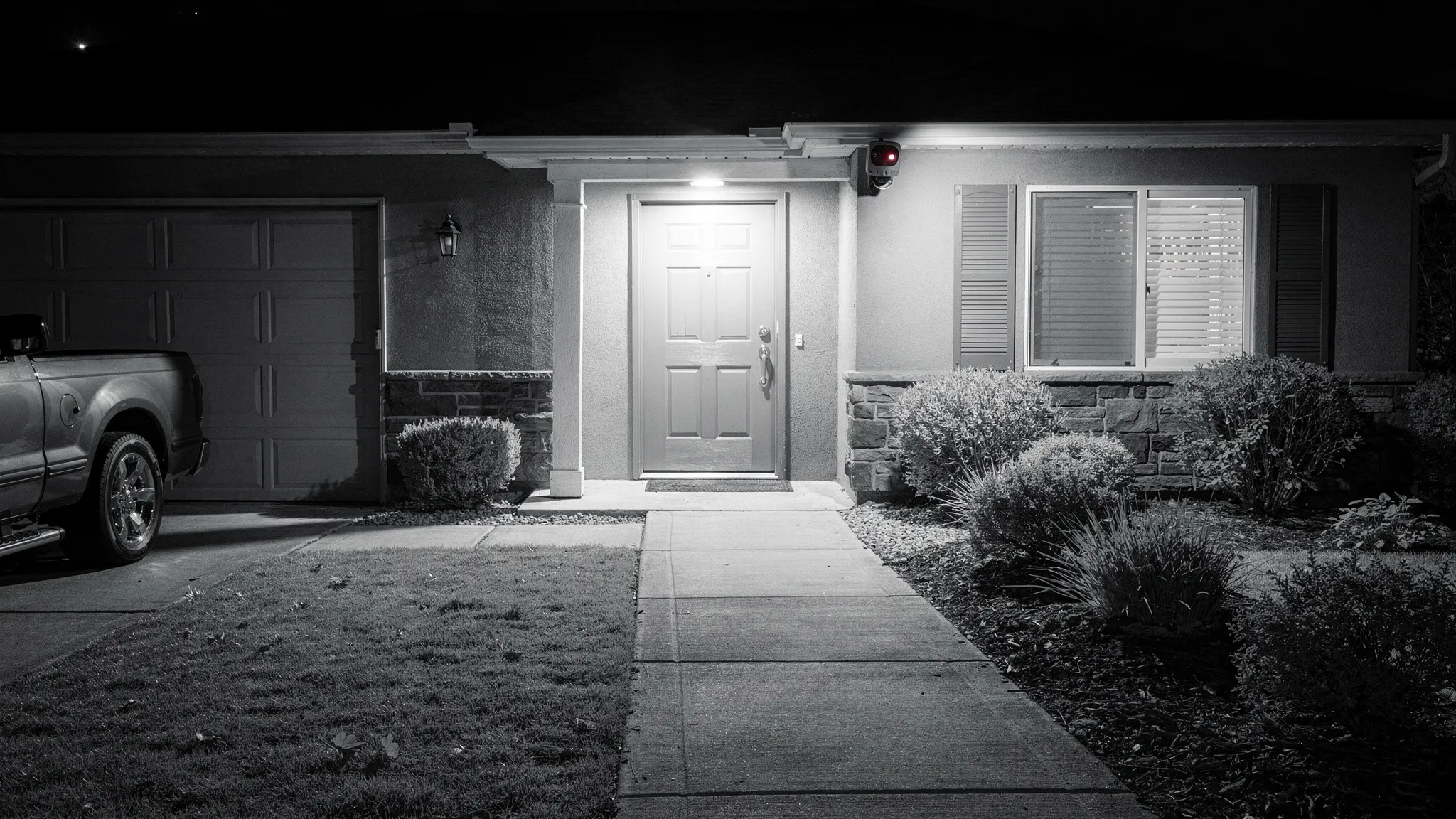 A home exterior at night with a security camera mounted under the eaves, showing an infrared-lit view of a front door and driveway area, with clear visibility of the entrance and path.